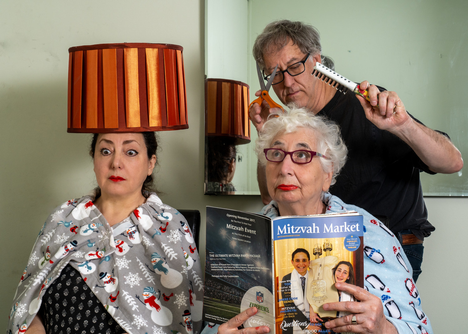 A man play-acting at giving two women a haircut, one with a magazine one with a lampshade