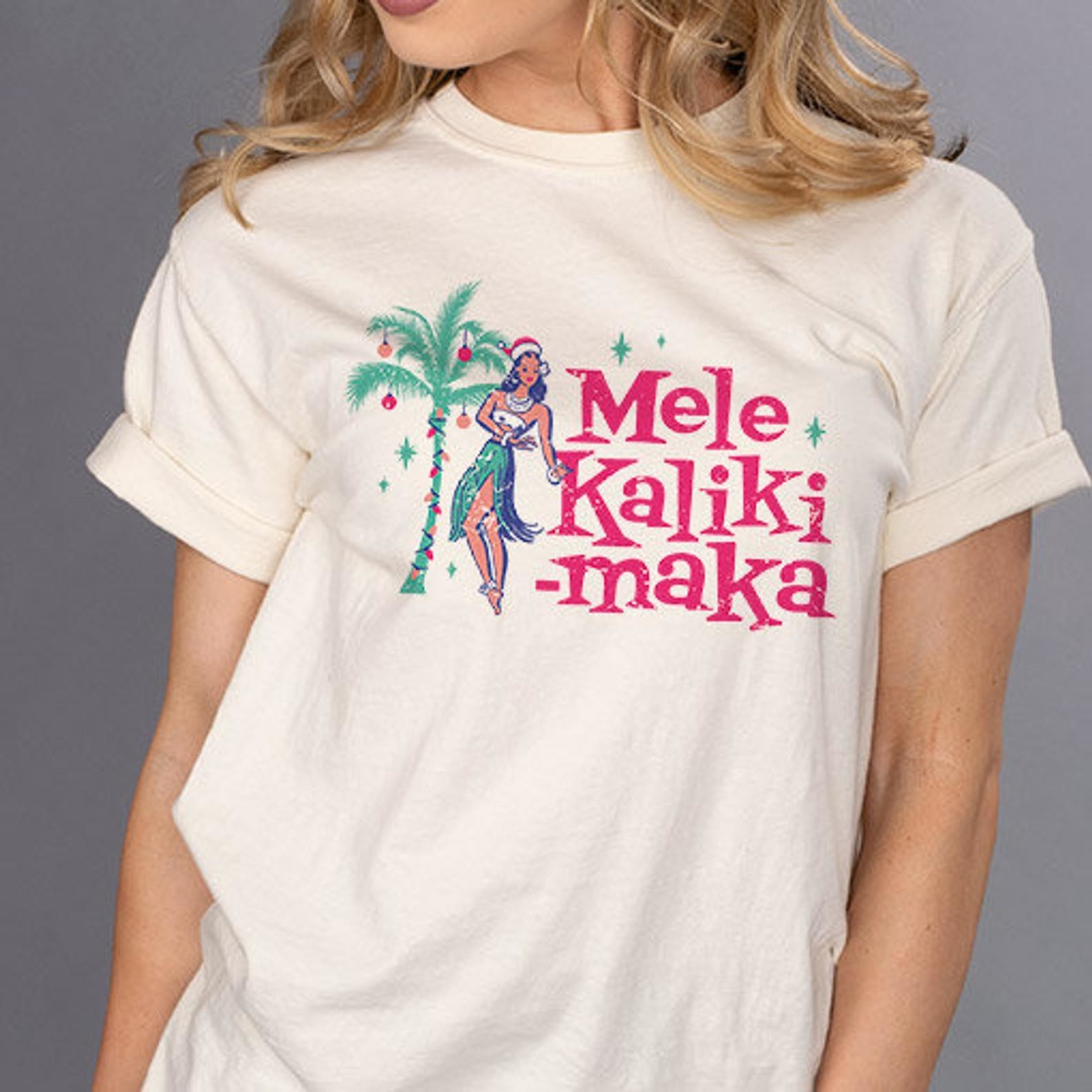 a white tee with a palm tree on it covered in ornaments, a hawaiian dancer, and &quot;mele kaliki-maka&quot; on it