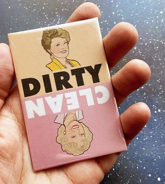 A hand holding a pink and tan magnet printed with a portrait of Blanche from Golden Girls with the word "Dirty" underneath, followed by the word "Clean" and a portrait of the character Rose oriented upside-down