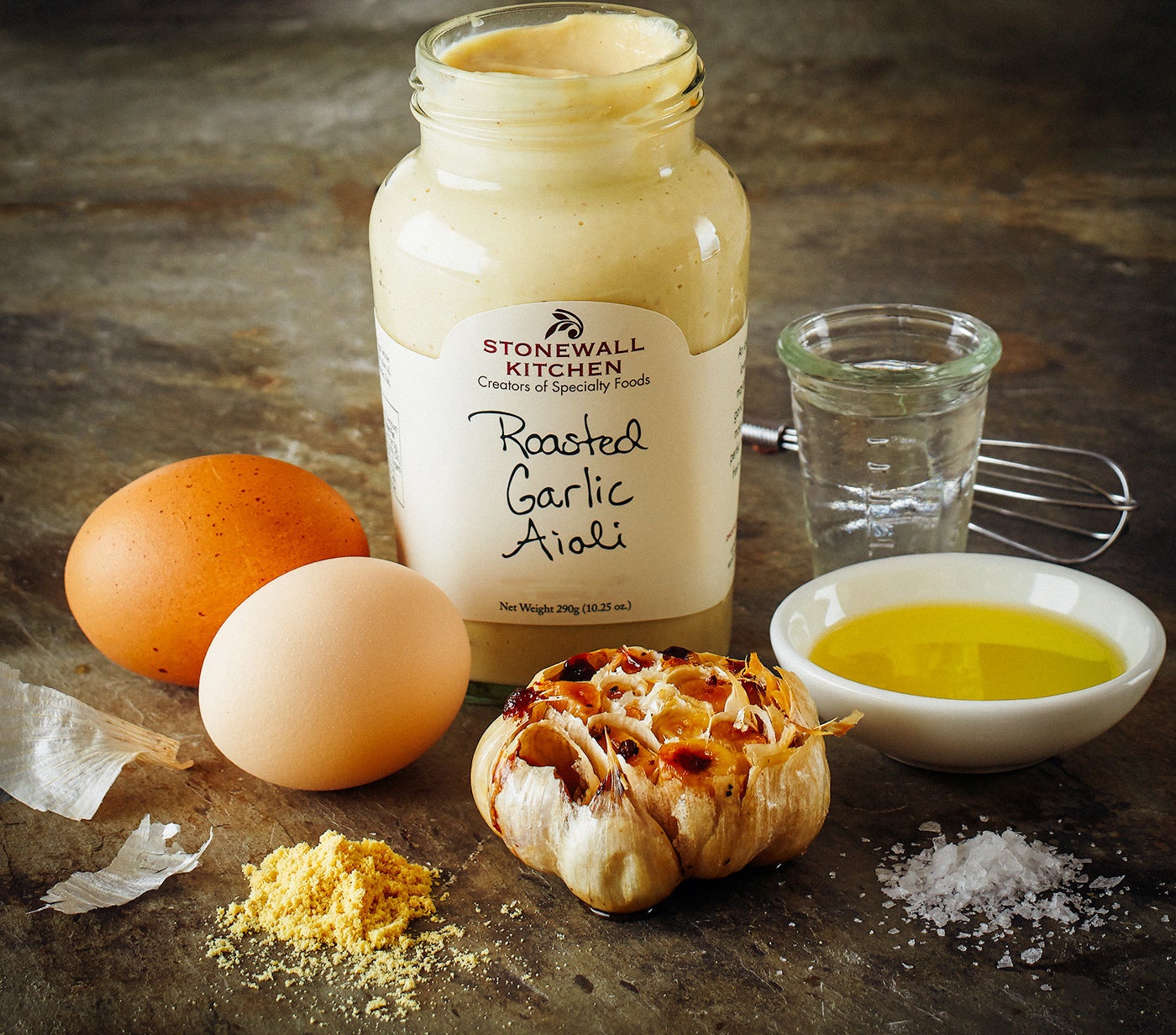 A jar of Stonewall Kitchen roasted garlic aioli surrounded by fresh garlic cloves, eggs, and oil on a countertop 