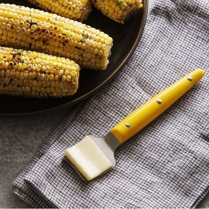 The butter knife with a curved head and yellow handle sitting next to a bowl of charred corn on the cob 