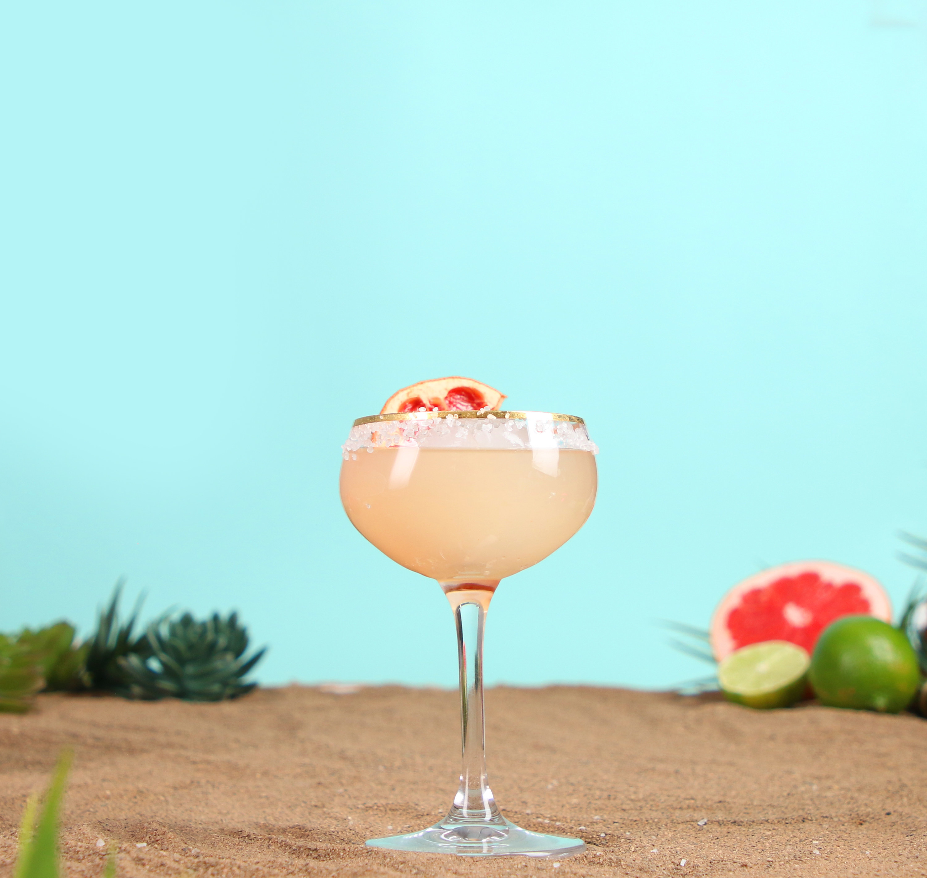 A colourful cocktail in front of a bright blue background on top of sand with a grapefruit peal in the drink.