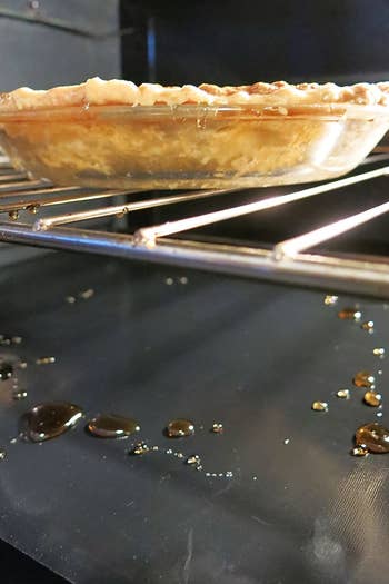 A pie baking in an oven lined at the bottom with one of the removable oven liners covered in drippings 