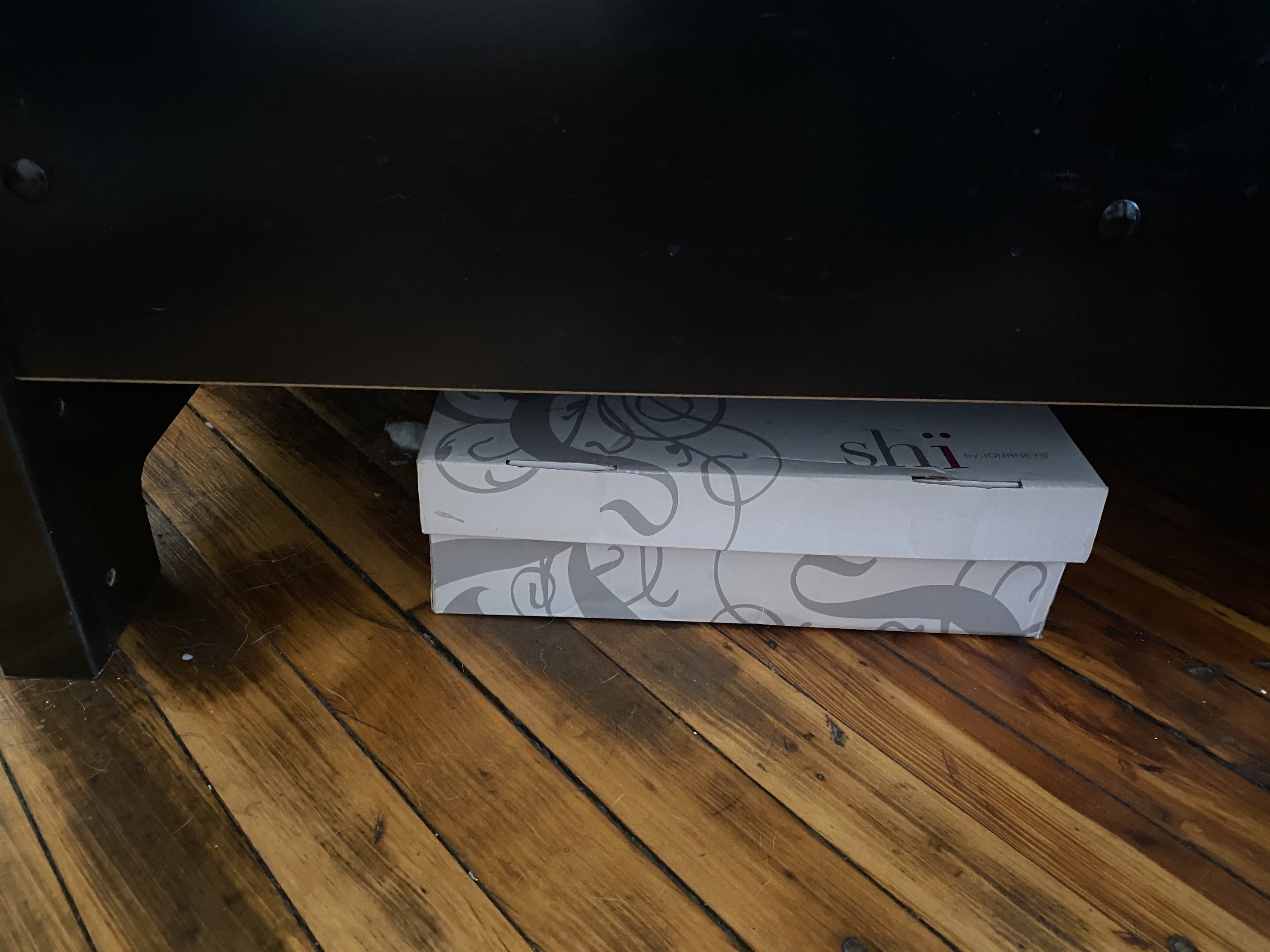 A white shoe box sits on a hardwood floor underneath a bed. 