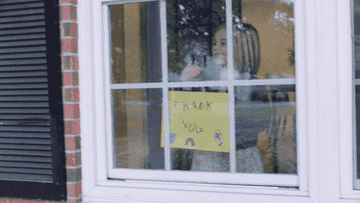 A young girl holds a sign that reads &quot;thank you&quot; and waves from inside her house 