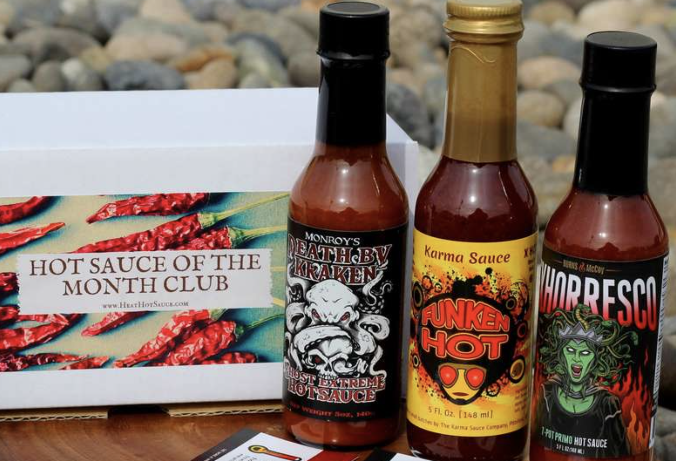 Three hot sauce bottles with colorful, quirky labels sit next to a hot sauce of the month club box with chile peppers on it