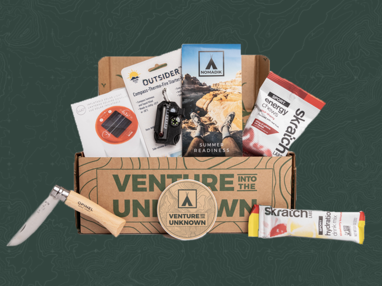 An open cardboard box with items like a camping knife, fire starter, energy chews, and more against a dark green background