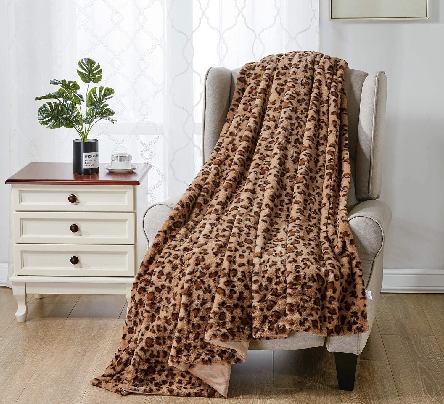 A leopard print blanket strewn over a cushioned chair next to a set of drawers that has a plant perched on top, for ambiance