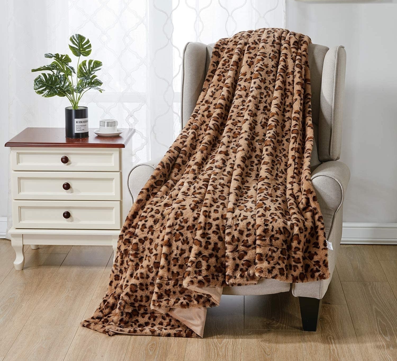 A leopard print blanket strewn over a cushioned chair next to a set of drawers that has a plant perched on top, for ambiance