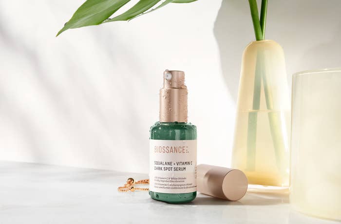 The Squalane and Vitamin C Dark Spot Serum in green bottle with a metallic pump top sitting on a table next to a vase