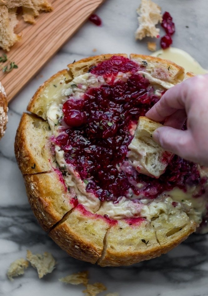 Someone dipping a piece of bread into a cranberry baked brie bread bowl.