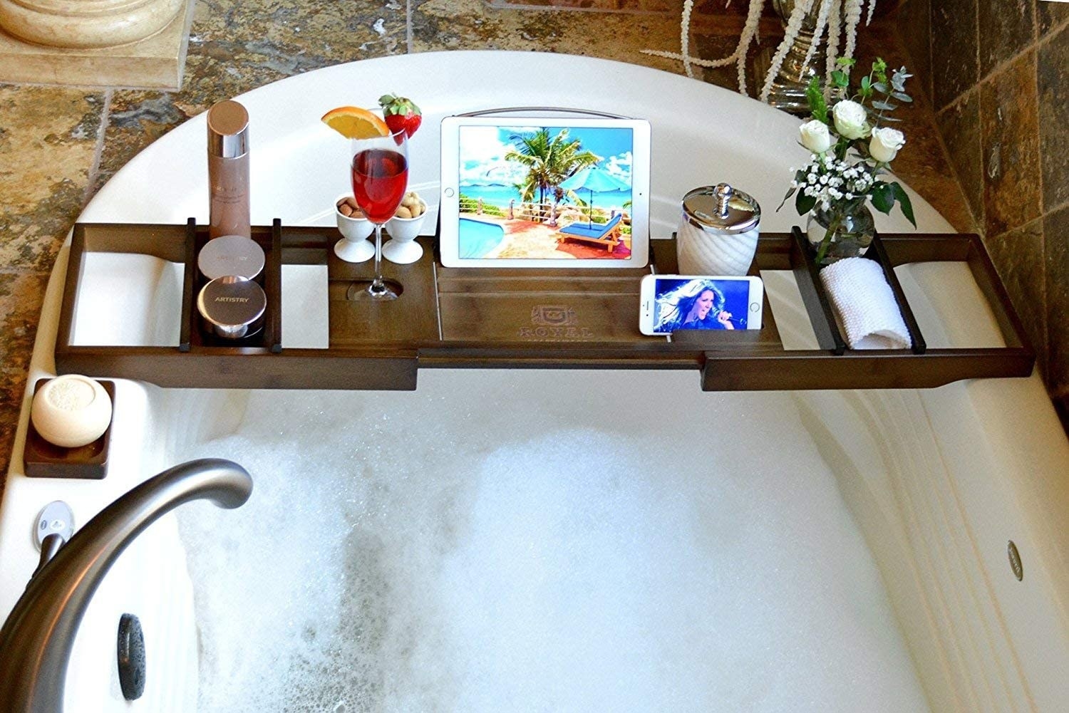 the bath caddy with wine, an iPad, a rose, and candles on top of it hung over a bathtub