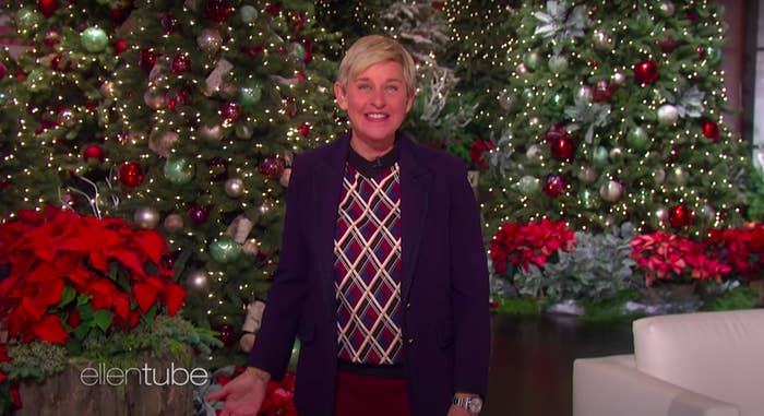 Ellen DeGeneres smiles and wears a festive holiday sweater and jacket on her talk show, standing in front of Christmas trees, decorated with lights and ornaments