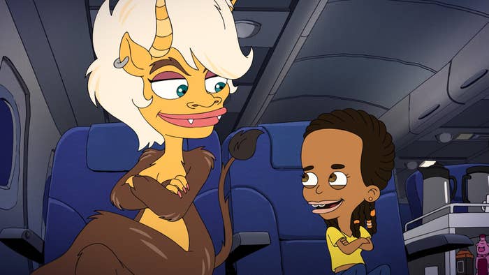 An animated monster, with big eyes, a pierced ear, and fangs, sits on an airplane beside a young Black girl, both of whom smile and have their arms crossed
