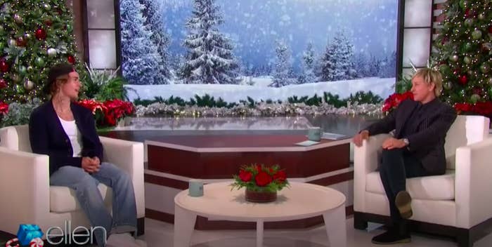 Justin Bieber, wearing jeans and a backward cap, sits in a white chair across from Ellen DeGeneres, wearing dark-colored clothing, on her talk show, which is decorated with Christmas trees and a wintry, snow-filled animation behind them