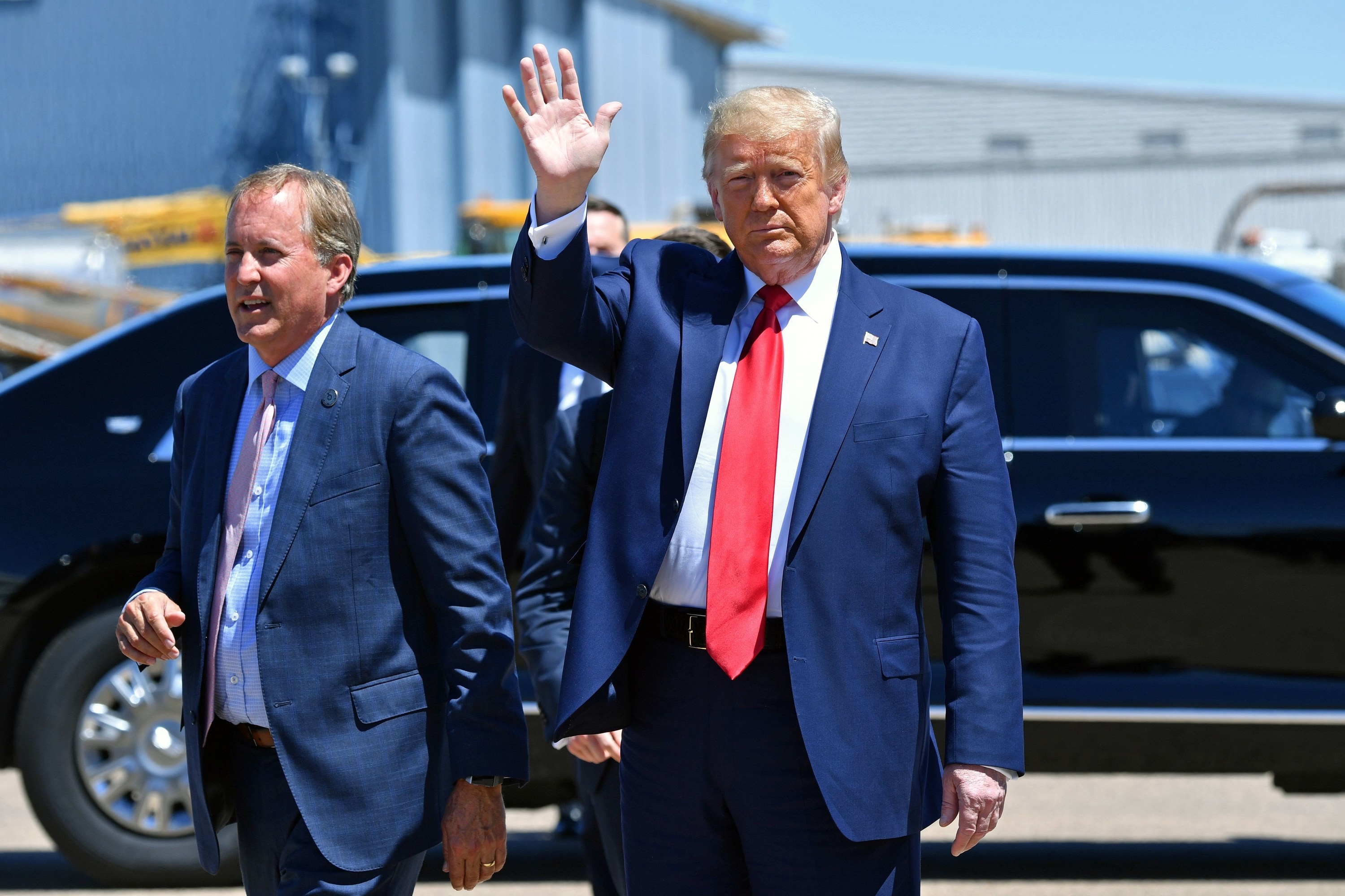 Donald Trump, wearing a big red tie, waves his hand, standing next to Ken Paxton outside in front of a black car from the presidential motorcade