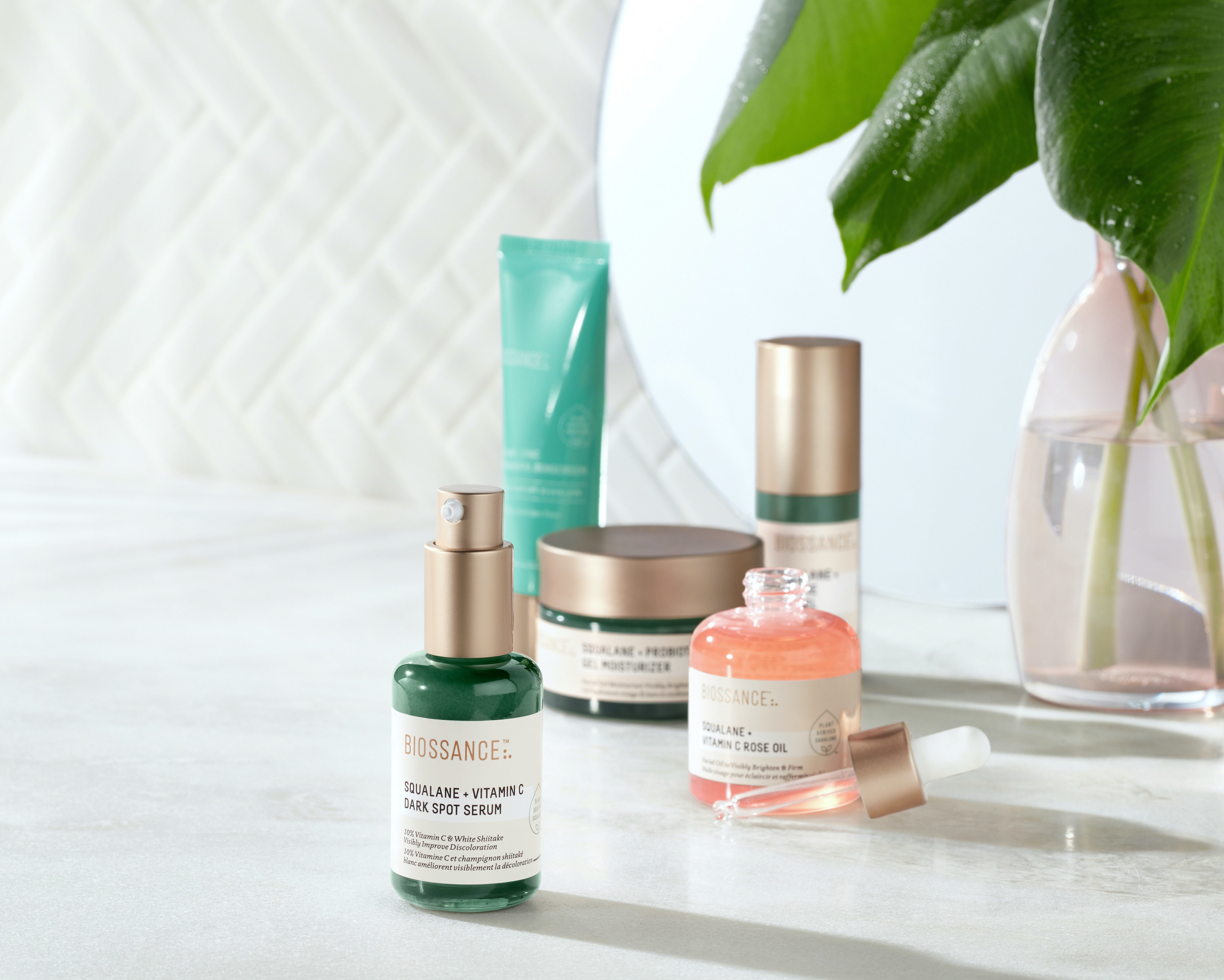 The Squalane and Vitamin C Dark Spot Serum in a green bottle with a metallic pump top sitting in front of a mix of other Biossance products 
