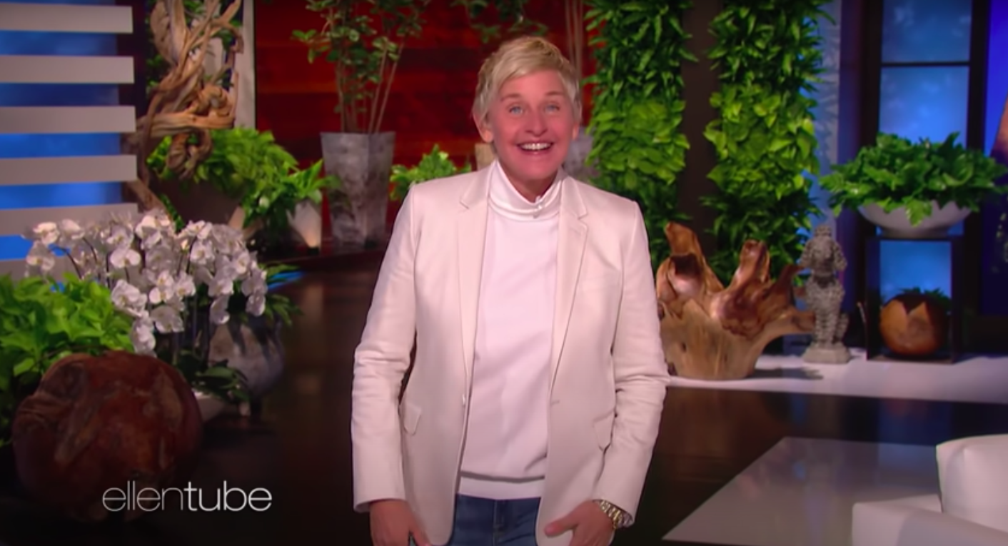 Ellen DeGeneres, wearing a white blazer and turtleneck, smiles widely at the camera