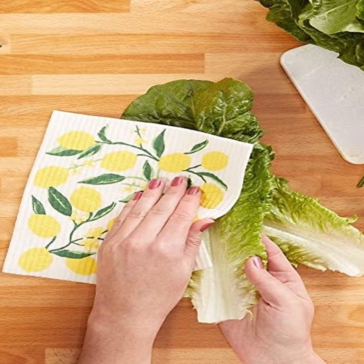 A hand wiping down a head of lettuce with a Swedish Dishcloth printed with lemons 