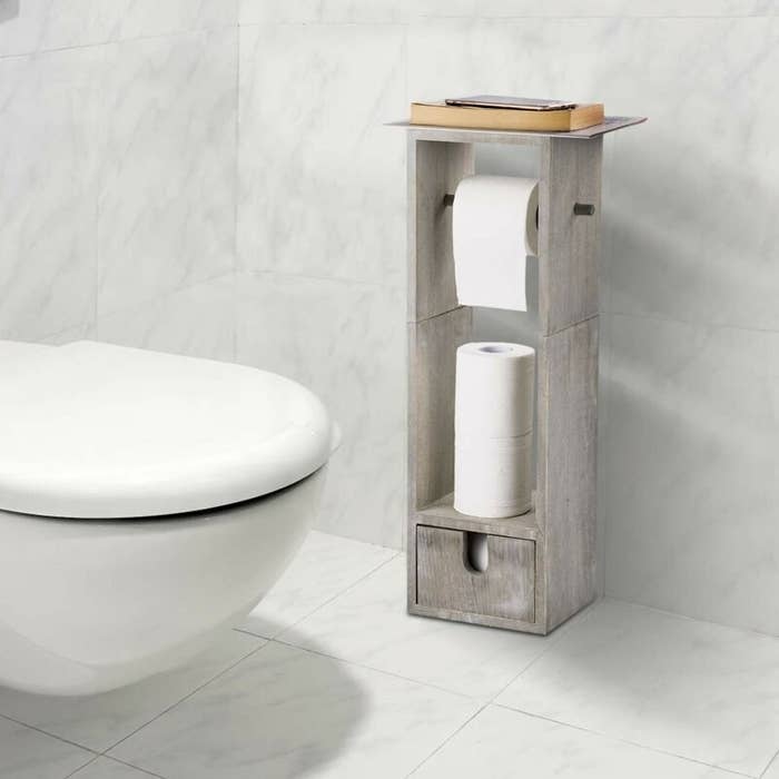 The toilet paper holder, which is long and rectangular, with a flat shelf on top, a rod for hanging the paper, a storage area to stack unused paper, and a small storage drawer at the bottom