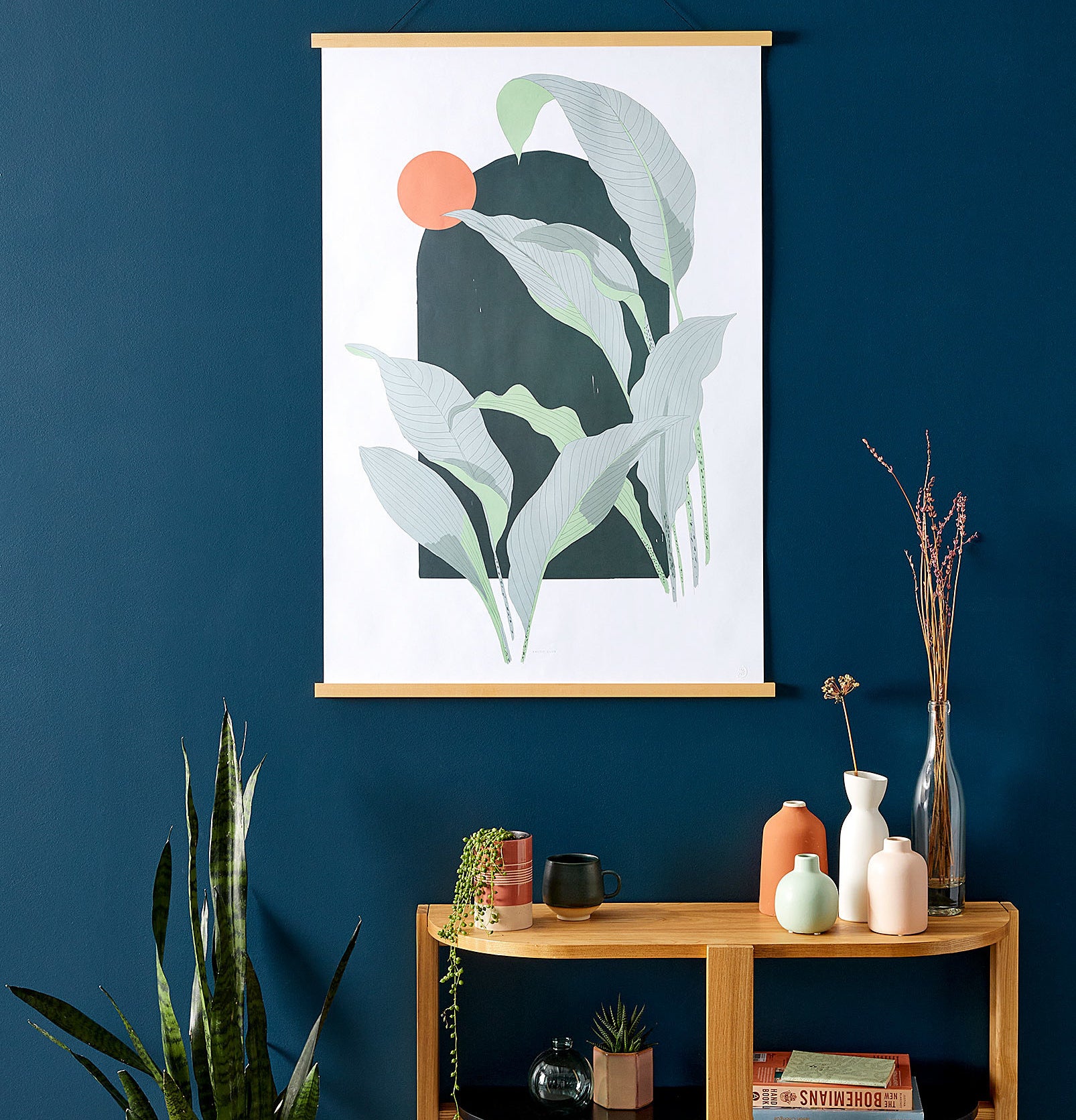A leafy art print hanging in a frame next to a snake plant and a side table with vases on it