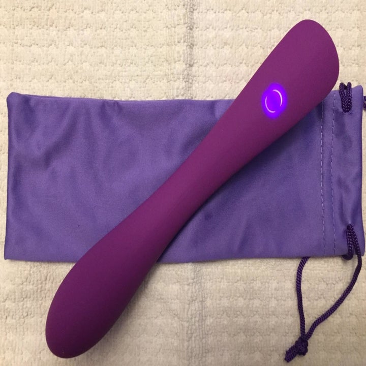 The slim, purple vibrating dildo with a glowing purple button sitting on top of the matching storage pouch 