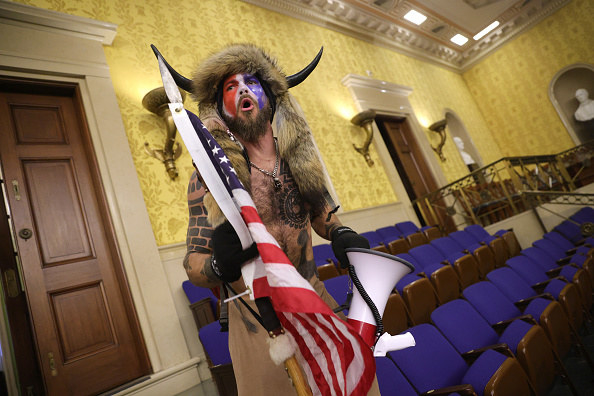 A shirtless man wearing horns and face yells as he stands inside the Capitol while holding the U.S. flag