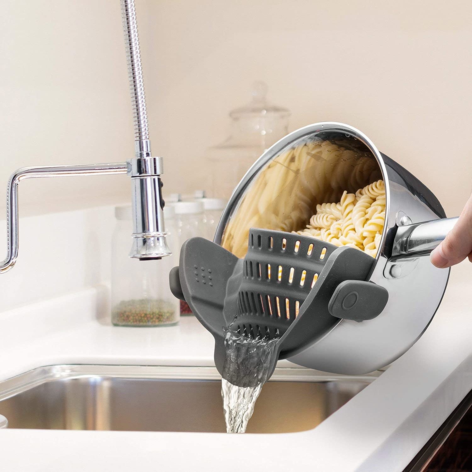 Model uses silver pot with clip-on black strainer to strain water out of a batch of pasta in a sink