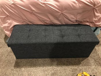 reviewer image of the tufted-top ottoman in dark grey in front of a bed