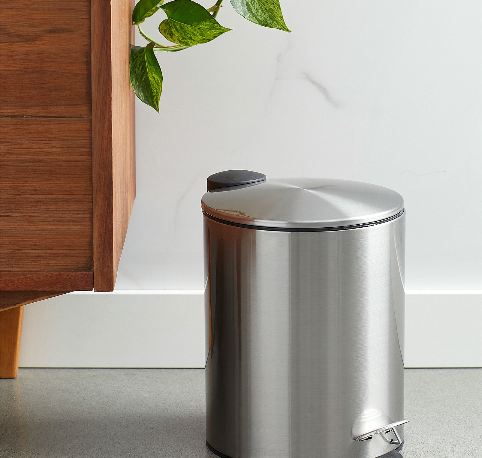 A metallic wastebasket with a pedal opening system next to a wooden dresser
