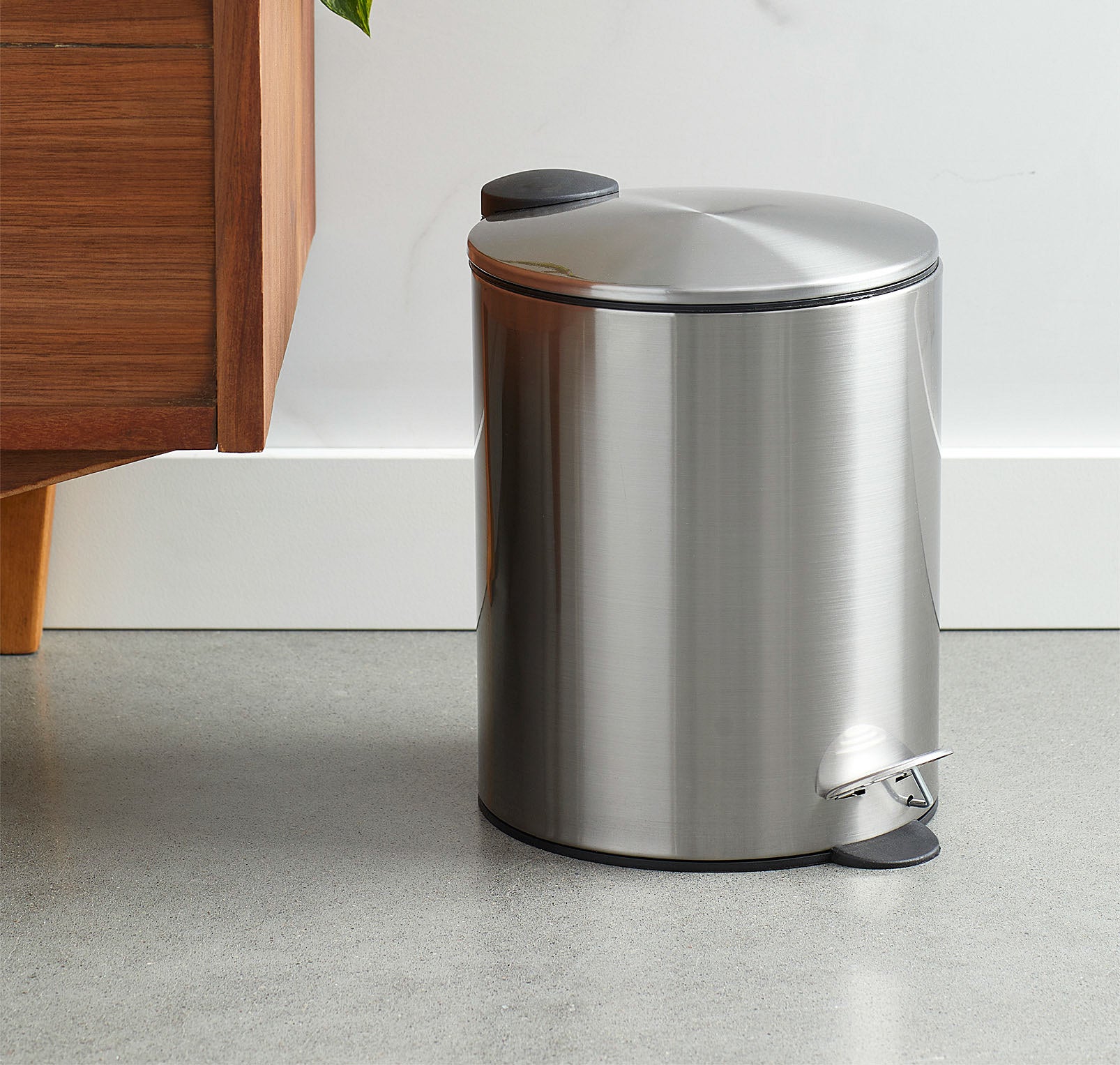 A metallic wastebasket with a pedal opening system next to a wooden dresser