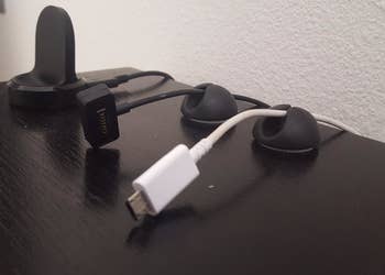 A review photo of three cable clips stuck to the top of a desk with three different cords stuck inside to keep them from slipping behind the desk 