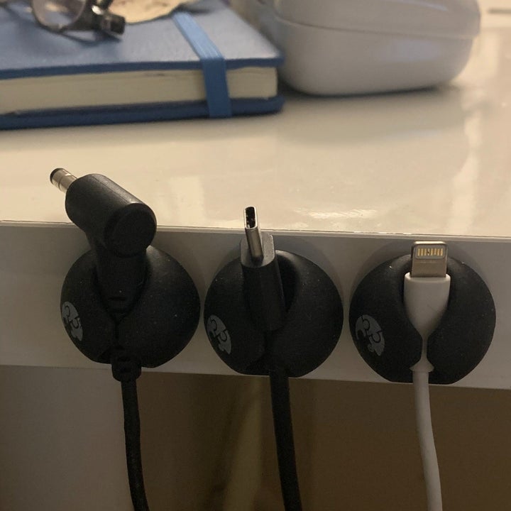A reviewer photo of three cable clips stuck to the side of a surface with three different charging cables secured inside to keep the cords from falling 