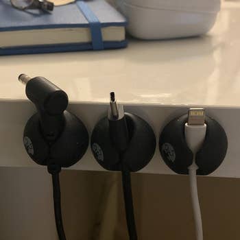 A reviewer photo of three cable clips stuck to the side of a surface with three different charging cables secured inside to keep the cords from falling 