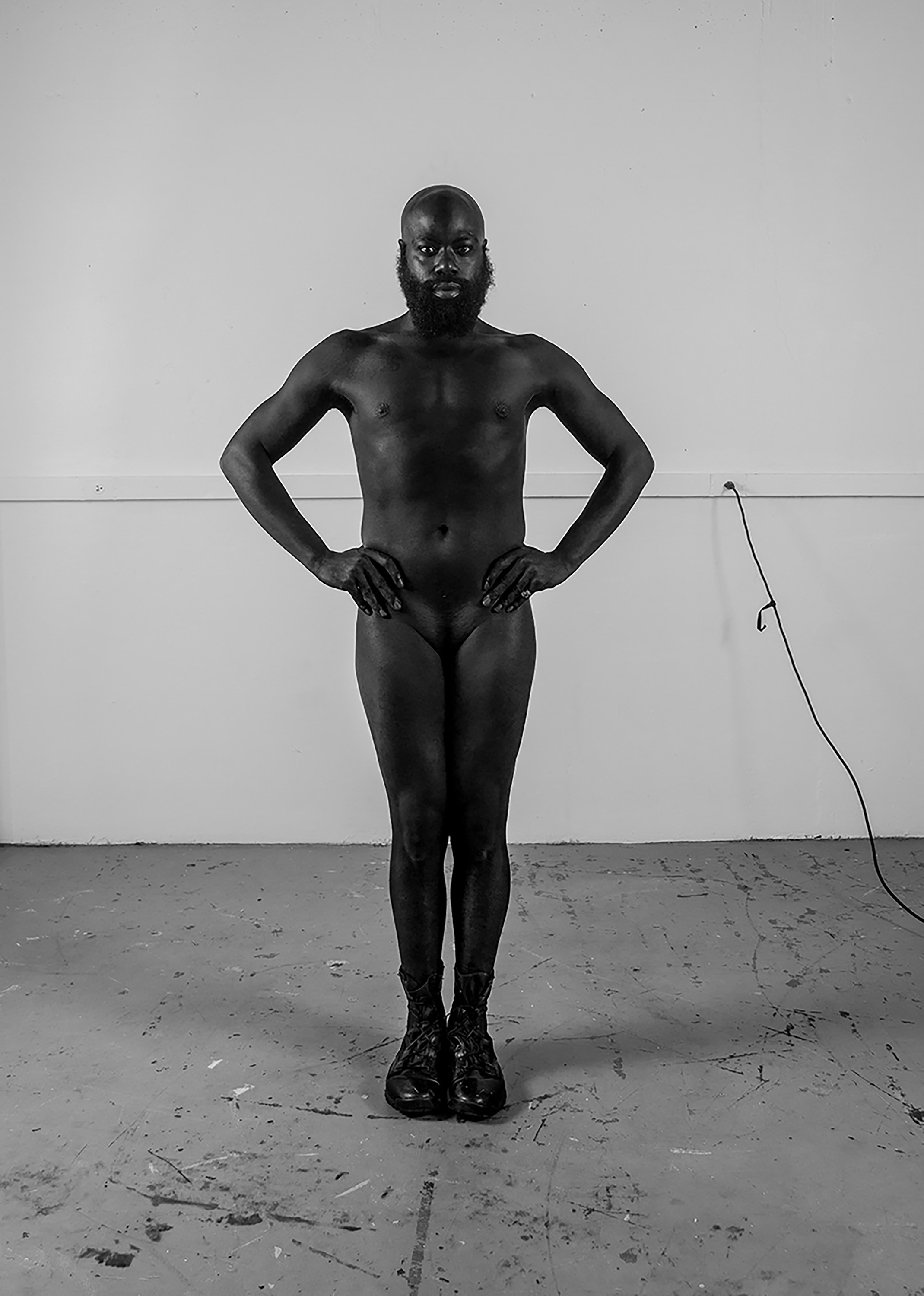 A man stands naked in a black-and-white portrait on a painted floor in black boots