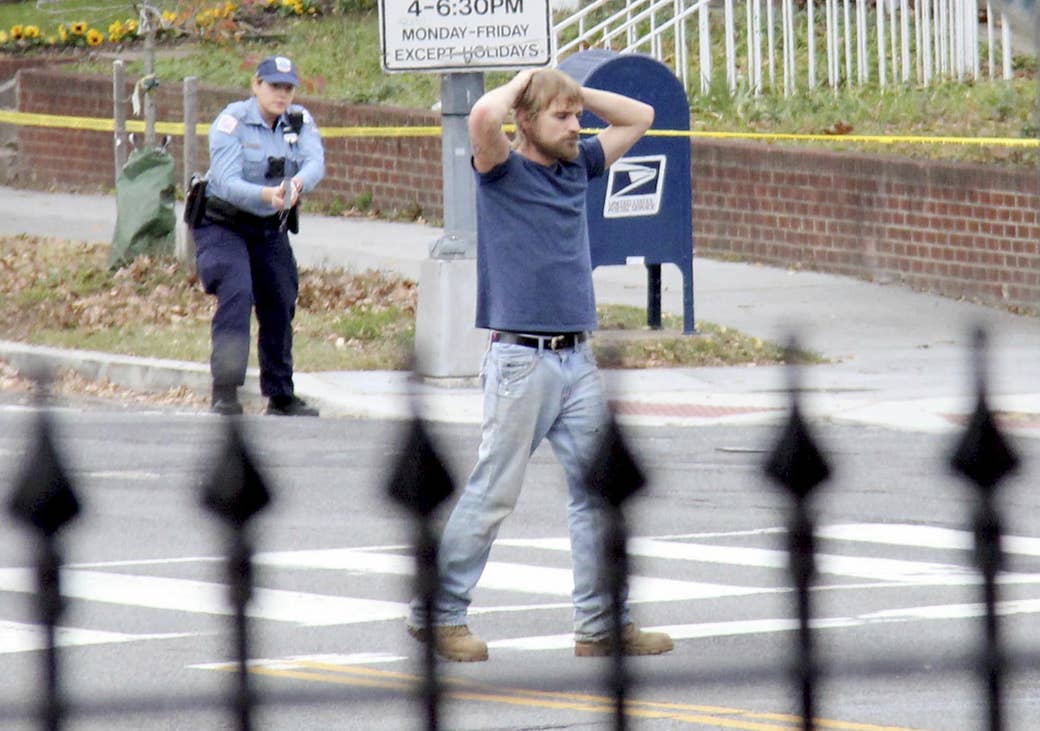 A white man stands at a crosswalk with his hands on his head while a police officer with her gun drawn stands several yards behind him