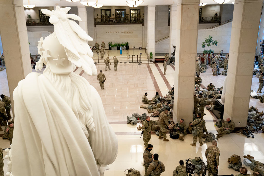 Armed National Guard members sitting and sleeping on the floor in front of a statue inside the Capitol