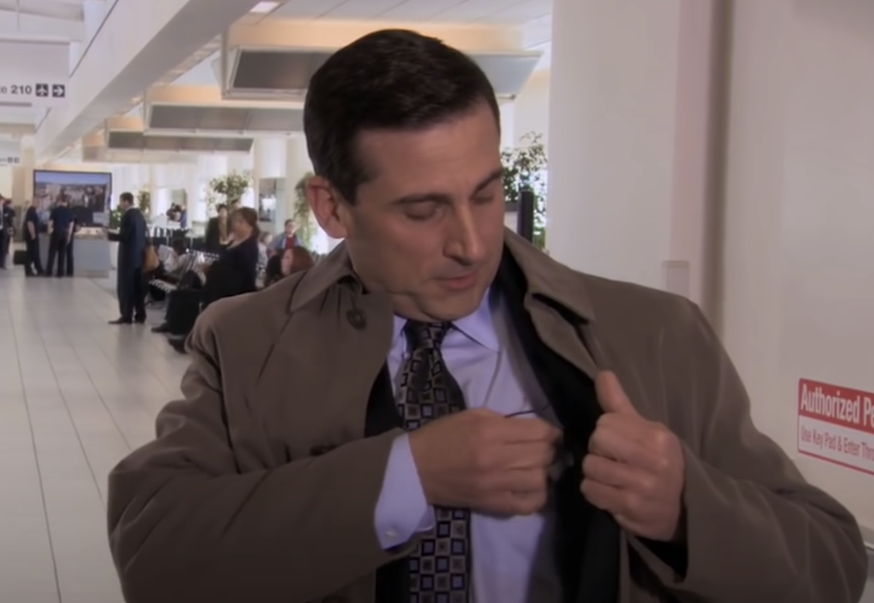 a man wearing a suit, tie, and trenchcoat stands in an airport, taking off a microphone