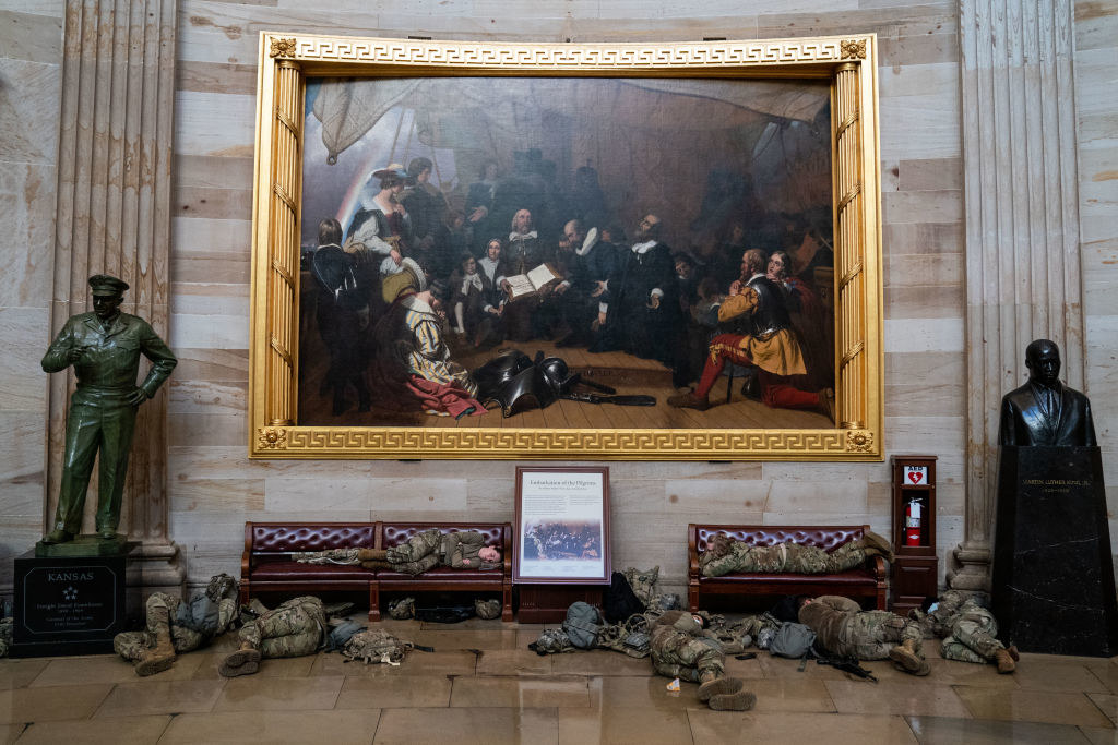 Two National Guard members lie on benches in front of a massive painting flanked by a bust and a statue inside the Capitol