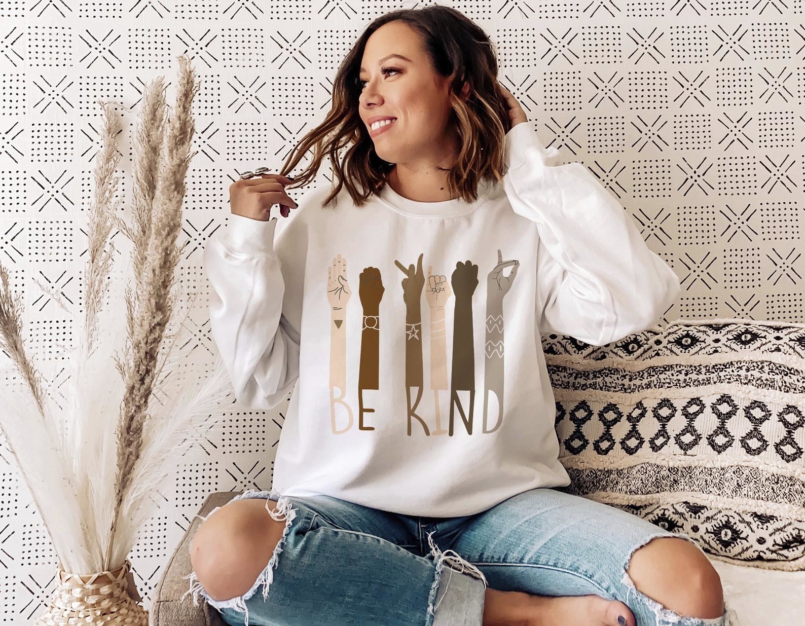 a model wearing the graphic crewneck sweatshirt in white that reads be kind and has different color arms signing each letter