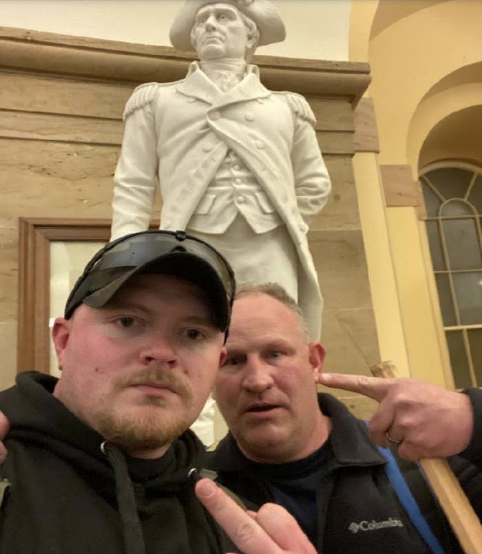 Two white men take a selfie in front of a statue, one flipping off the camera and the other pointing at him