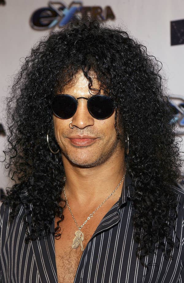 Slash smiling at the camera wearing his signature sunglasses, with his hair down and in a button-up top