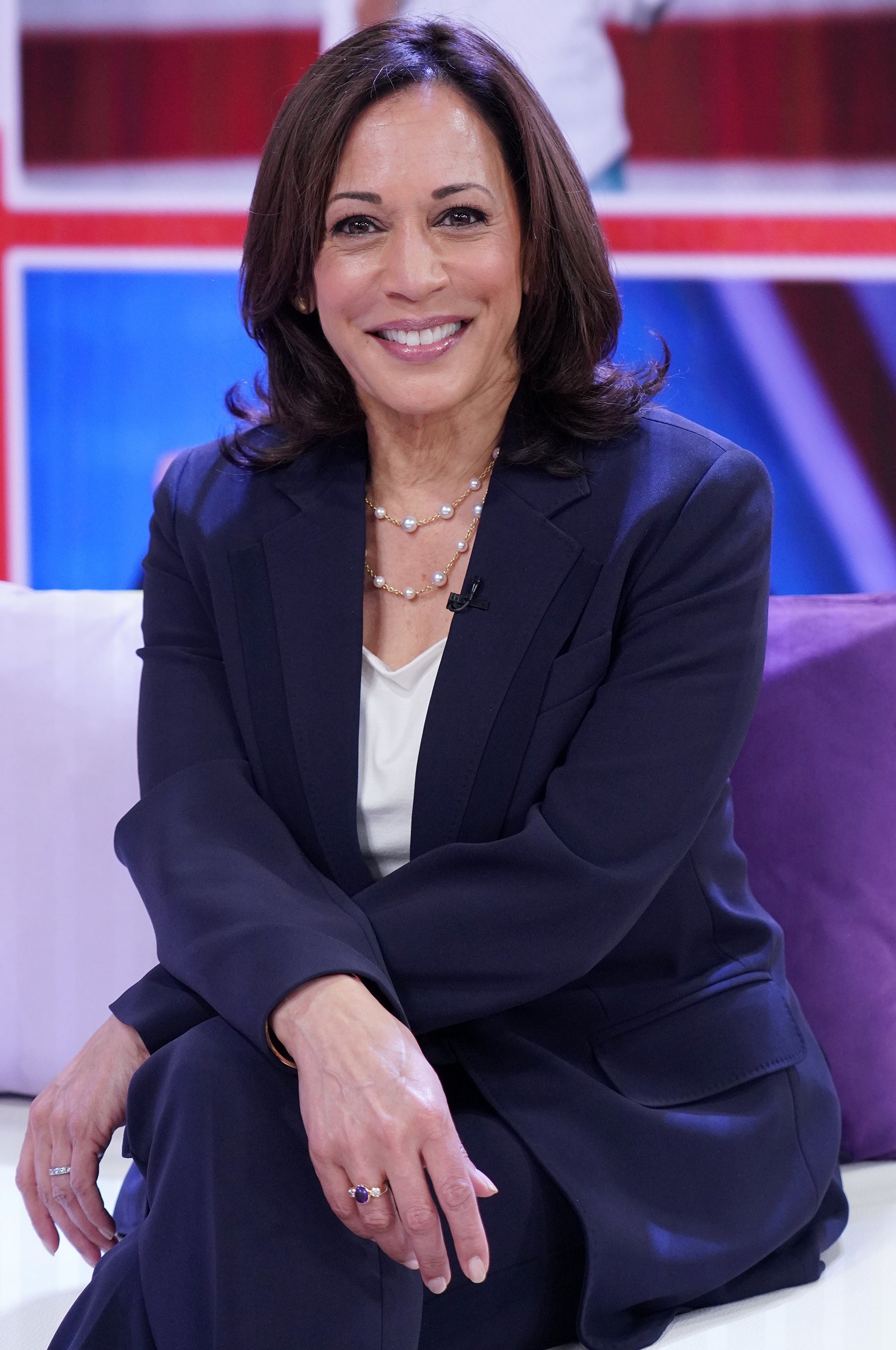 Kamala Harris smiling as she sits on a talk show couch in a navy blue suit with a white shirt