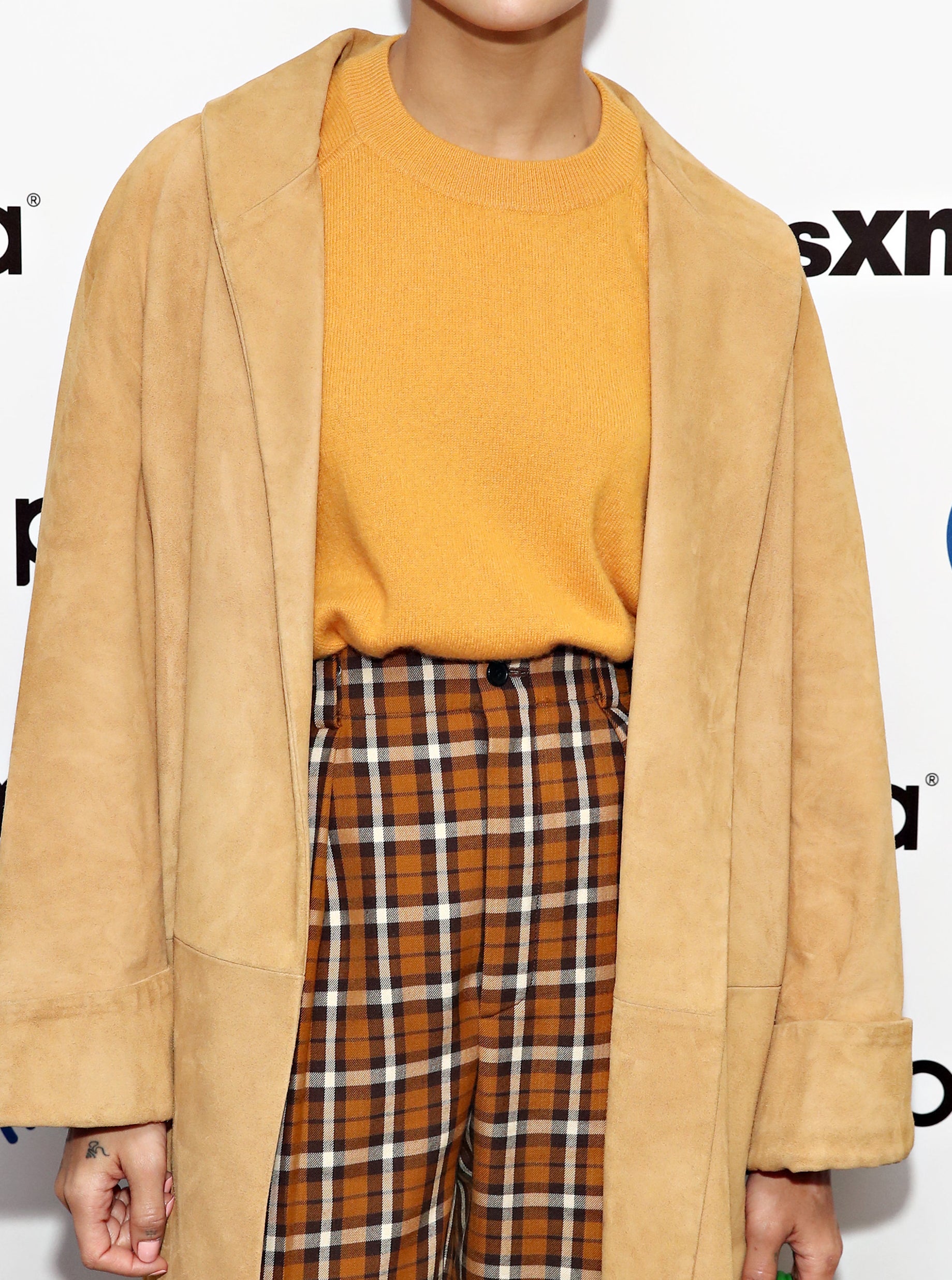 Zoe Kravitz at a red carpet event, with a pixie cut, wearing an orange sweater, plaid bands, and a coat