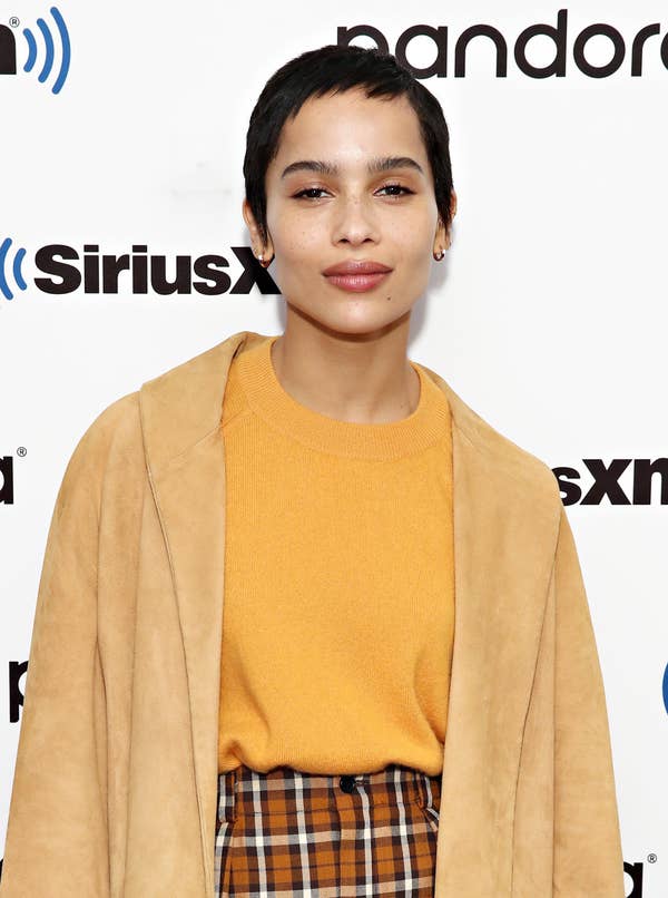 Zoe Kravitz at a red carpet event, with a pixie cut, wearing an orange sweater, plaid bands, and a coat