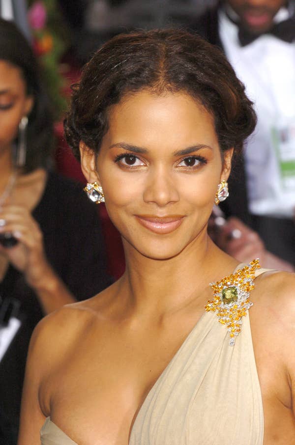 Halle Berry smiling, wearing a single shoulder toga-esque dress with a brooch and her hair pulled back