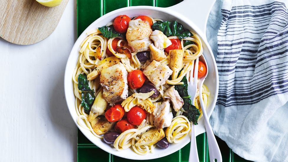 Top down image of chunks of barramundi sitting on a bed of linguine with kale, cherry tomatoes, olives and artichokes