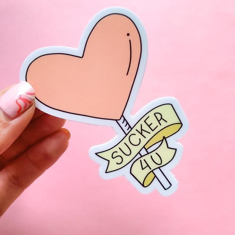 Hand holding the sticker shaped like a heart-shaped lollipop with a ribbon around it with the text &quot;Sucker 4U&quot;