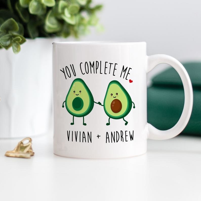 The white mug with two halves of an Avocado holding hands with the text "You complete me" and the couple's name on the bottom