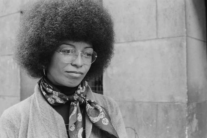 A woman with an afro and glasses stands in front of a stone building. 